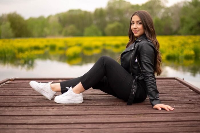 Fiatal nő fekete bőrdzsekiben és fehér sportcipőben egy fa stégen ül, a Vekeri-tó és sárga virágok látványos háttérrel, Debrecen közelében készült portréfotó.