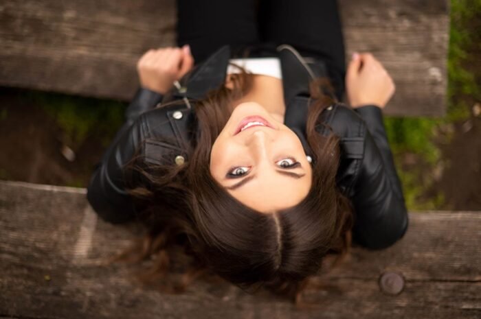 Fiatal nő fekete bőrdzsekiben háton fekve, felfelé nézve mosolyog egy fa padon, kreatív kültéri portréfotózás során.