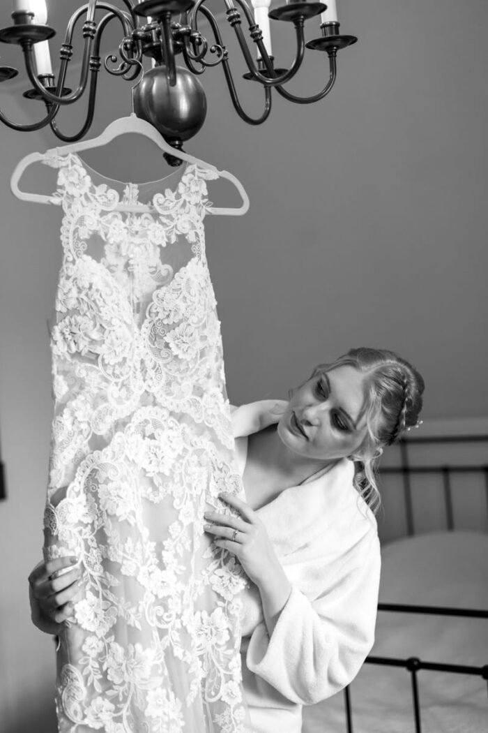 A menyasszony gyengéden tartja kezében az esküvői ruháját, miközben a csipkés részleteket csodálja. A kép fekete-fehér, amely kiemeli a ruha finom mintáit és a pillanat meghittségét.