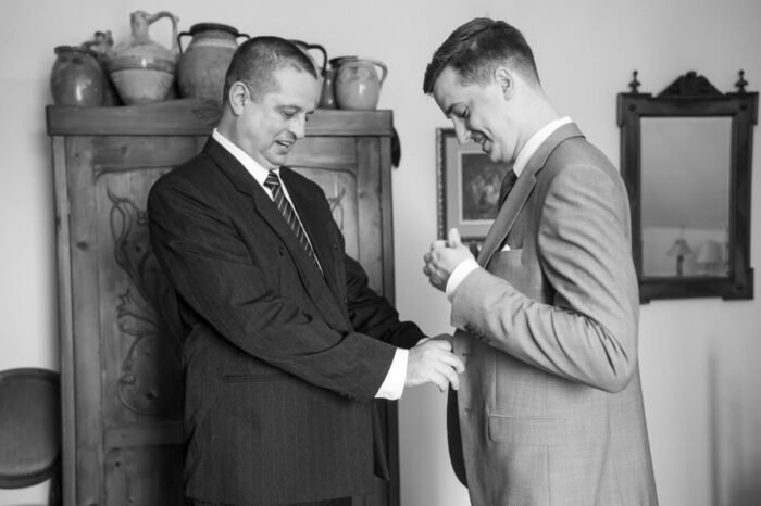 Édesapja segít a vőlegénynek az öltöny gombjának igazításában. A háttérben rusztikus fa szekrény és hagyományos tárgyak láthatók. A jelenet bensőséges, a készülődés pillanatait örökíti meg.