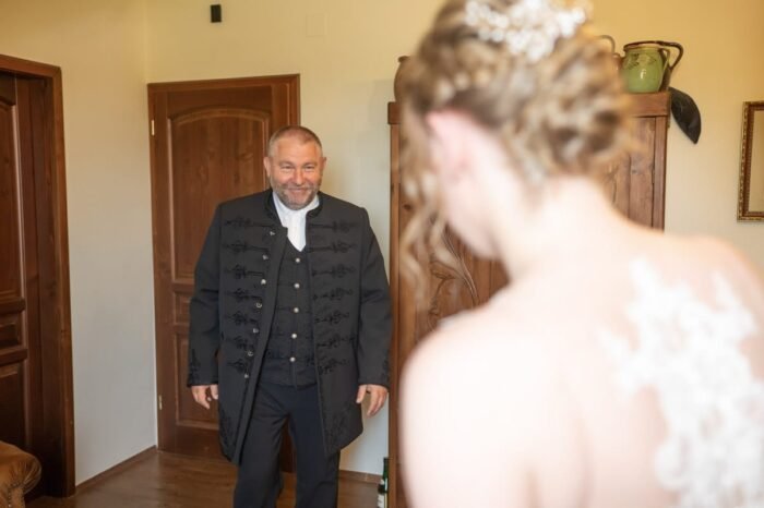 Az apa büszkén és szeretettel néz menyasszony lányára, miközben egy tradicionális, fekete díszmagyar öltözetet visel. A menyasszony háttal áll, ruhájának csipkés hátkivágása látható. A háttérben rusztikus ajtók és bútordarabok, melyek otthonos légkört teremtenek.