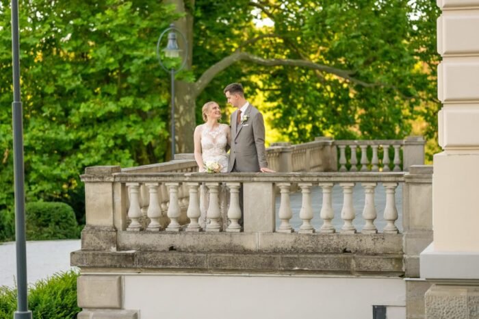 Egy boldog menyasszony és vőlegény áll egy elegáns erkélyen, őket körülvevő buja zöld lombok között. A menyasszony csipkés menyasszonyi ruhában, a vőlegény szürke öltönyben mosolyogva néznek egymásra. A klasszikus oszlopos erkély és a természet harmóniája egy romantikus hangulatot teremt.