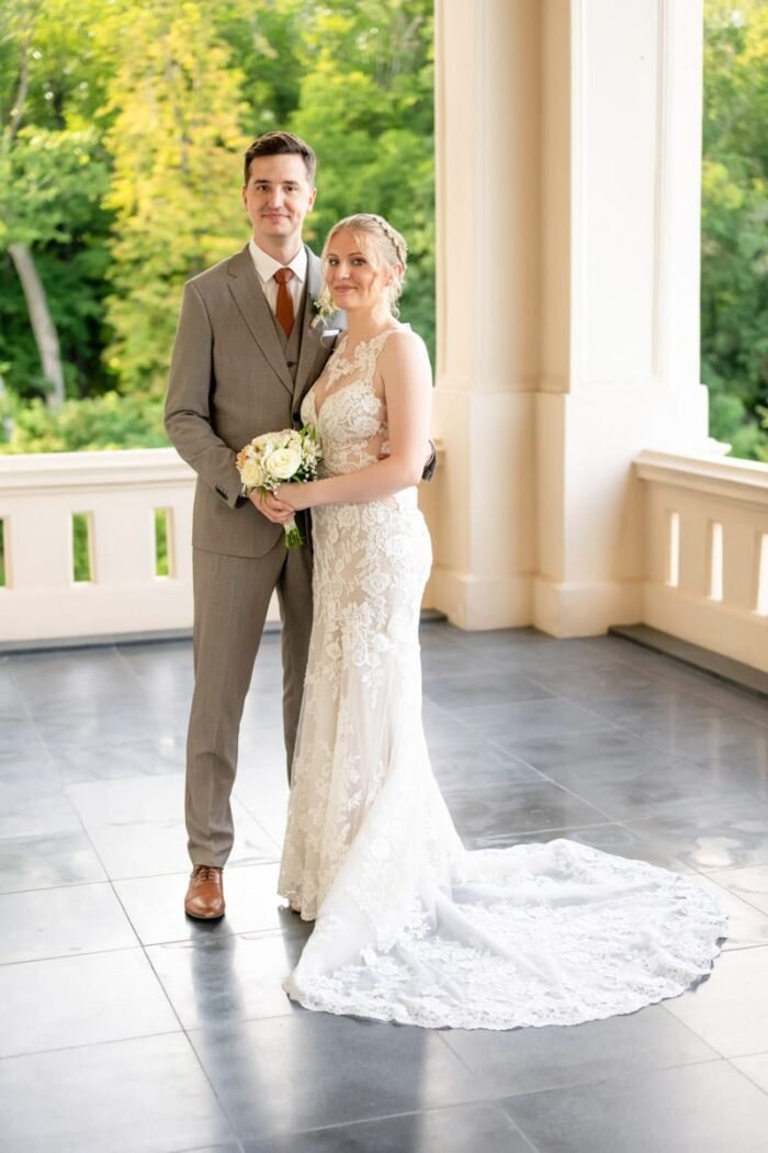 Az ifjú pár szorosan egymás mellett áll egy elegáns terasz boltívei között. A menyasszony csipkés, uszályos ruhában tartja virágcsokrát, miközben finoman mosolyog. A vőlegény szürke öltönyt és narancsszínű nyakkendőt visel, magabiztosan állva. A háttérben zöldellő fák emelik ki a jelenet természetes szépségét.