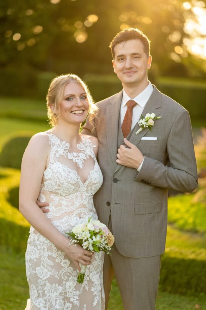 Az ifjú pár szorosan egymás mellett áll egy gyönyörű parkban, naplemente fényében. A menyasszony csipkés ruhájában virágcsokrot tart, mosolya boldogságot sugároz. A vőlegény világosszürke öltönyt és narancs árnyalatú nyakkendőt visel, kezét elegánsan a zakóján tartva. A háttérben zöld növényzet és lágy, aranyló fények teszik teljessé a pillanatot.