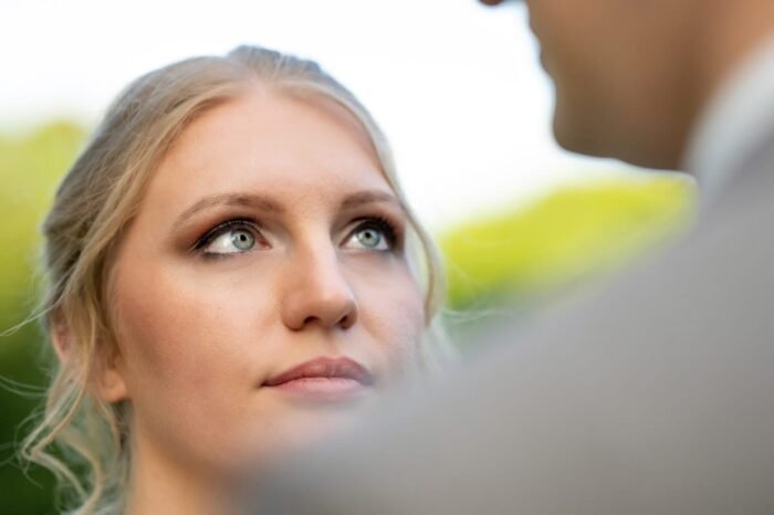 Egy meghitt pillanatban az ara szemeinek finom részletei kerülnek fókuszba, miközben tekintetét a vőlegény felé irányítja. A háttérben lágy zöld árnyalatok teszik romantikussá a hangulatot.