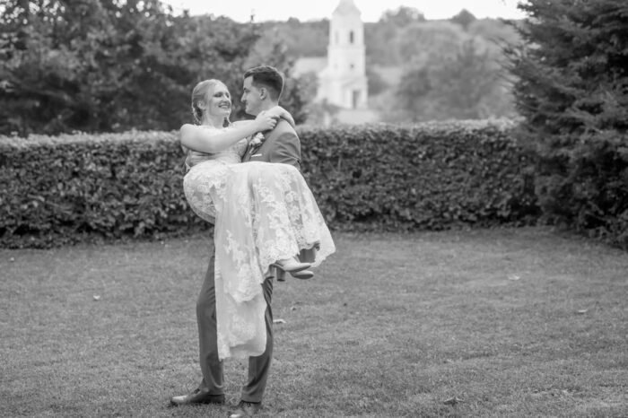 A vőlegény boldogan tartja karjaiban a menyasszonyt egy gyönyörű kerti háttér előtt, miközben mindketten mosolyognak, a távolban egy templom tornya is látható.