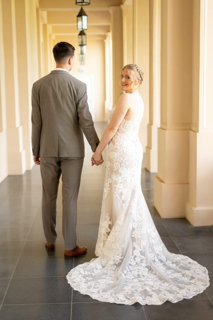 A menyasszony és a vőlegény kéz a kézben sétálnak egy elegáns kastély folyosóján, a menyasszony hátrafordulva mosolyog a kamerába, megmutatva gyönyörű csipkés ruhájának részleteit.