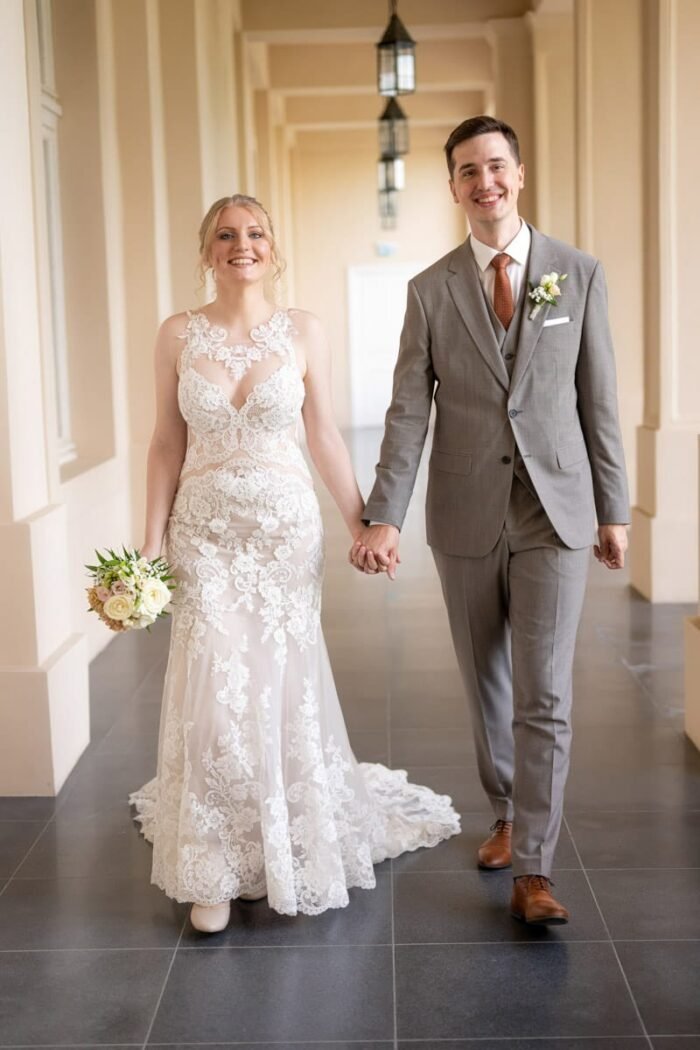 Az ifjú pár kéz a kézben, mosolyogva sétál egy elegáns kastély világos folyosóján, a menyasszony csipkés ruhát és virágcsokrot tart, a vőlegény elegáns öltönyt visel.