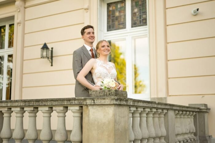 Az ifjú pár mosolyogva áll a füzérradványi kastély elegáns erkélyén, a menyasszony csipkés ruhát és virágcsokrot tart, míg a vőlegény szeretetteljesen öleli át.