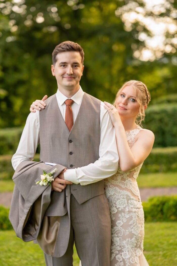 Az ifjú pár mosolyogva pózol egy zöldellő parkban, a menyasszony gyengéden a vőlegény vállára teszi a kezét, míg a vőlegény elegánsan öltönyben áll.