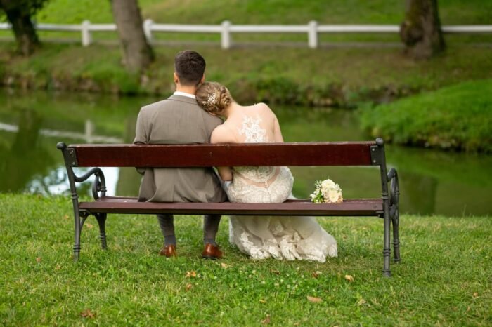 Az ifjú pár hátulról látható, ahogy egy tóparti padon ülnek, a menyasszony a vőlegény vállára hajtja fejét, mellettük a virágcsokor. Nyugodt és romantikus pillanat.