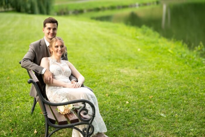 Az ifjú pár egy romantikus tóparti padon ül, mosolyogva és egymáshoz bújva, a háttérben zöldellő fű és a tó látható.