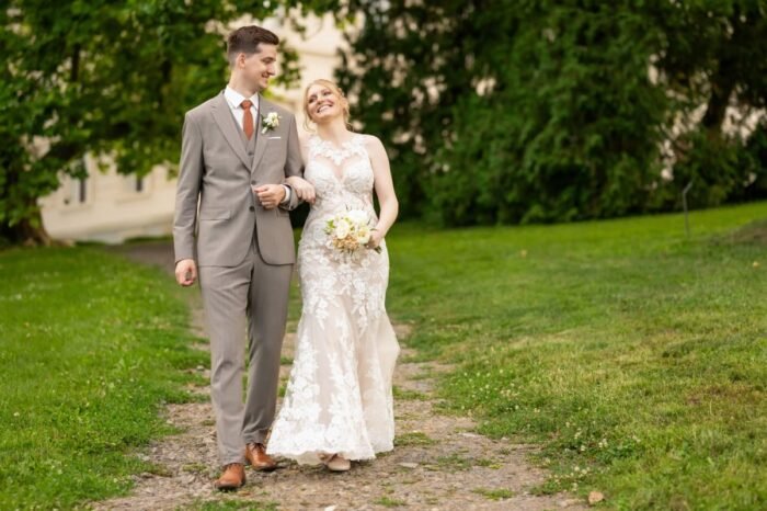 Az ifjú pár mosolyogva sétál kéz a kézben egy zöldellő parkban, a menyasszony virágcsokrot tart, míg a vőlegény szeretetteljesen néz rá.