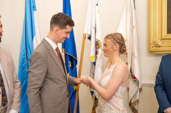 A menyasszony és a vőlegény mosolyogva néznek egymásra, miközben a gyűrűhúzás meghitt pillanatát élik át egy polgári szertartás során, zászlókkal a háttérben.