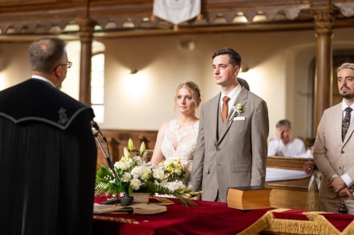 Az ifjú pár komolyan és meghatottan áll az oltárnál egy egyházi szertartáson, virágdíszítés és szimbolikus tárgyak között. A lelkész a szertartást vezeti.