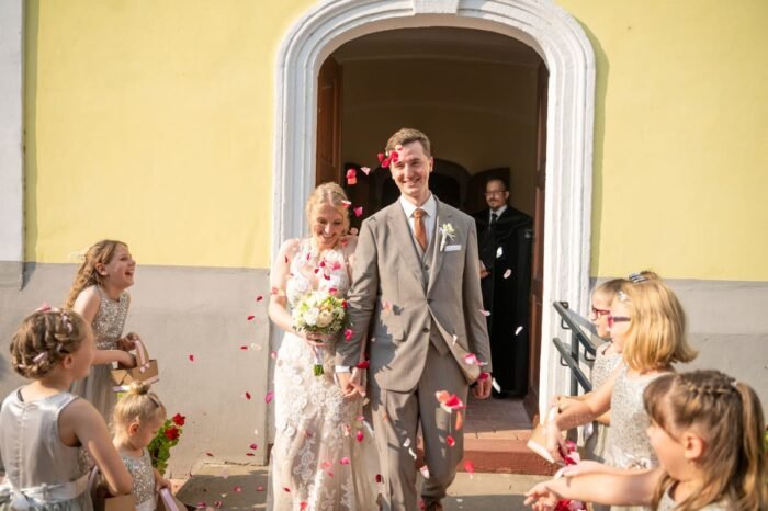 Az ifjú pár mosolyogva lép ki a templomból, miközben gyerekek rózsaszirmokat szórnak rájuk, és vidám hangulat uralkodik a pillanatban.