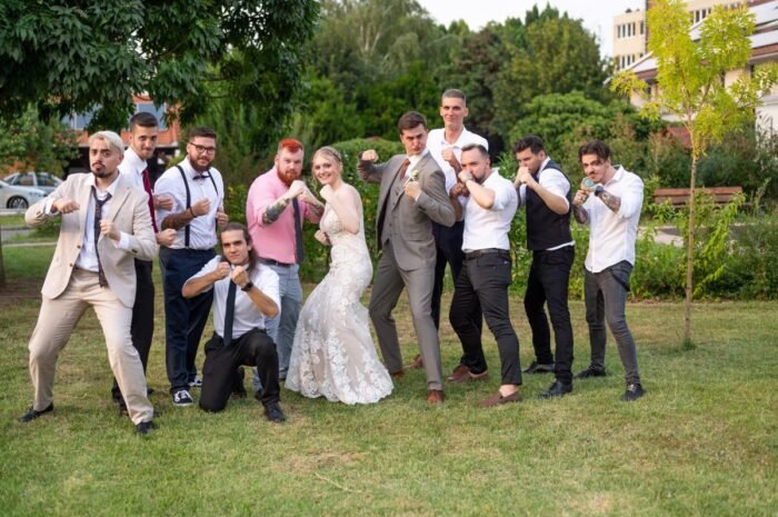 Az ifjú pár és barátaik harcias pózban állnak a zöldellő kertben, vidám és játékos hangulatú esküvői fotón.