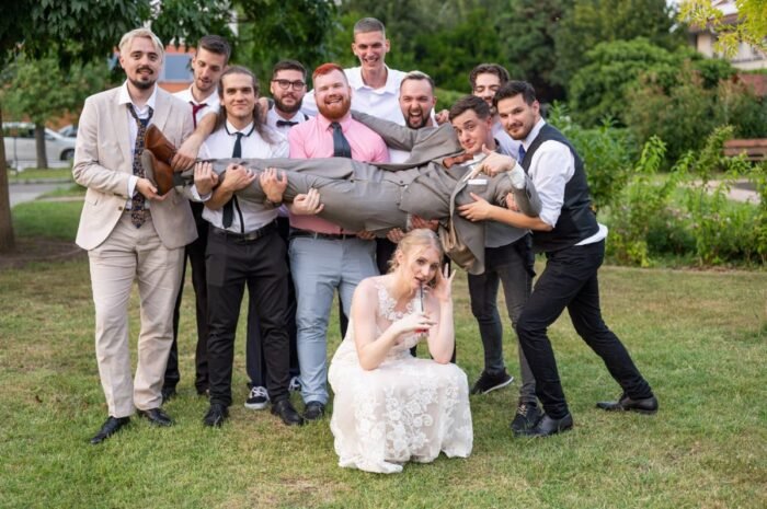 A vőlegényt barátai felemelik a levegőbe, miközben a menyasszony humorosan pózol a földön ülve, italával a kezében. Vidám hangulatú esküvői kép zöldellő környezetben.