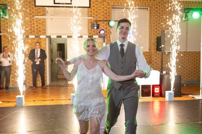 Az ifjú pár mosolyogva táncol a nyitótáncon, miközben pirotechnikai elemek emelik az esemény hangulatát. A háttérben vendégek tapsolnak és figyelik őket.