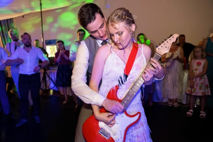 Egy esküvői fotón a pár gitározik együtt a táncparketten, miközben a vendégek mosolyogva nézik őket.