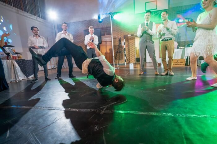 A férfi a táncparketten mozdulatokkal lenyűgözi a közönséget, miközben a vendégek tapsolnak és szórakoznak. A háttérben a zöld fények világítanak, és a menyasszony az előző táncpartnerével együtt élvezi a bulit. Az esküvői hangulat tele van élettel és jókedvvel, miközben a barátok és a családtagok is hozzájárulnak az esemény szórakoztató hangulatához.