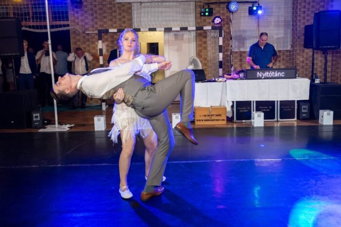 A menyasszony és a vőlegény dinamikusan táncolnak a parketten, miközben a vőlegény hátra hajolva van a menyasszony által tartva. A háttérben a DJ pult látható, ahol a zenét keverik, és a "Nyitótánc" feliratú kijelző világít. Színes fények világítják meg a táncparkettet, miközben a pár élvezi az első táncot.