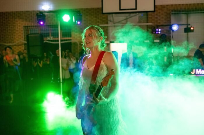 A menyasszony a színpadon ragyog, miközben a zöld füstfelhőben és színes fények között játszik. A táncoló násznép a háttérben élvezi a zenét, miközben ő maga a figyelem középpontjában áll, flitteres ruhájában és gitárjával.