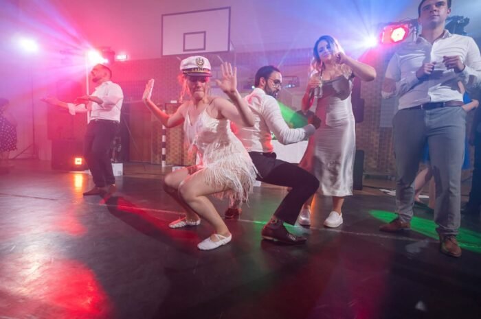 A násznép tagjai szórakoztató táncot lejtenek a színes fények között, miközben az egyik nő egy kapitányi sapkában és flitteres ruhában vezeti a mozdulatokat. A háttérben a többi vendég is élvezi a táncot, és az egész terem tele van energikus mozdulatokkal és boldog pillanatokkal.