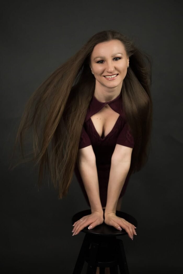 Portré fotózás Debrecen.Egy mosolygós portréfotó, amelyen egy bordó ruhás, hosszú barna hajú nő dinamikus pózban támaszkodik egy székre, miközben haja szabadon lebeg a sötét háttér előtt.