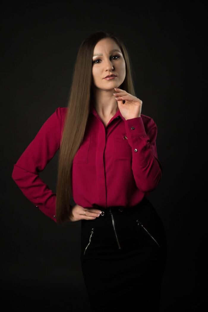 Debreceni portré fotózás - Egy elegáns portréfotó, amelyen egy hosszú barna hajú nő pink inget és fekete szoknyát viselve áll, kezét állához emelve, miközben sötét háttér előtt pózol.