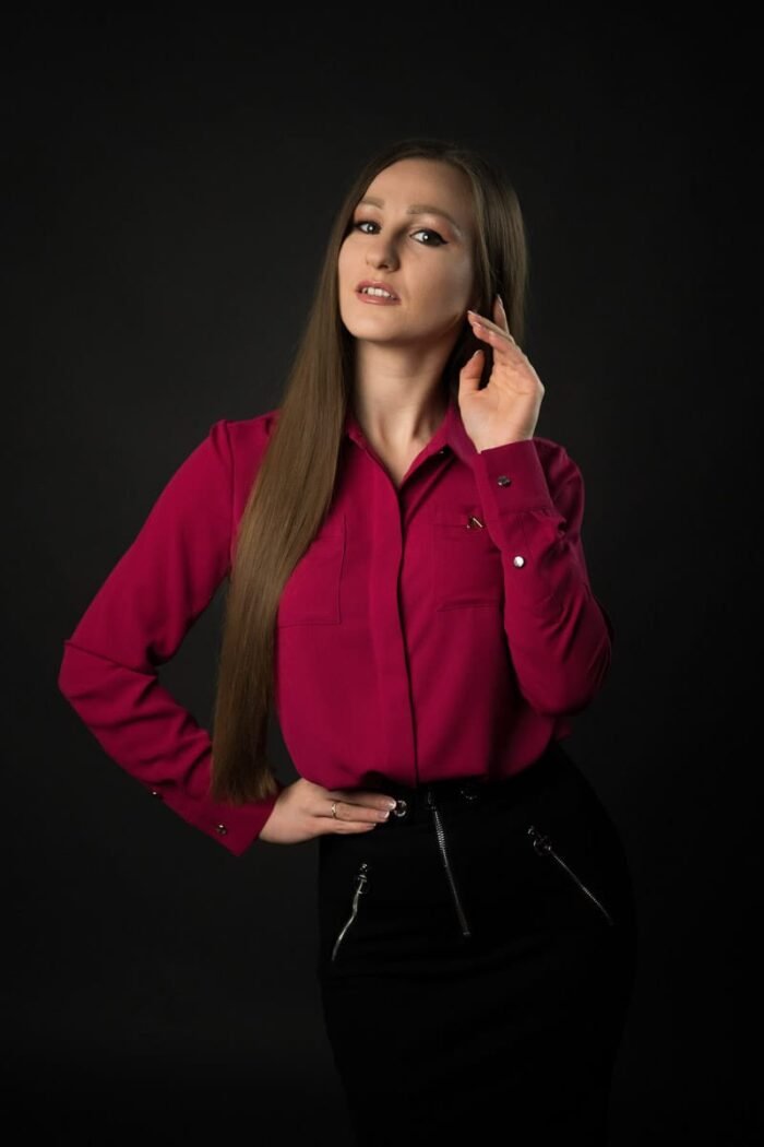 Portréfotó egy stúdióban, ahol egy hosszú barna hajú nő pink inget és fekete szoknyát viselve elegánsan pózol Debrecenben, sötét háttér előtt.