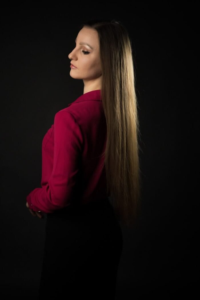 Portré fotózás Debrecenben.Portréfotó oldalnézetből, amelyen egy hosszú barna hajú nő pink inget és fekete szoknyát viselve elegáns pózban áll sötét háttér előtt.