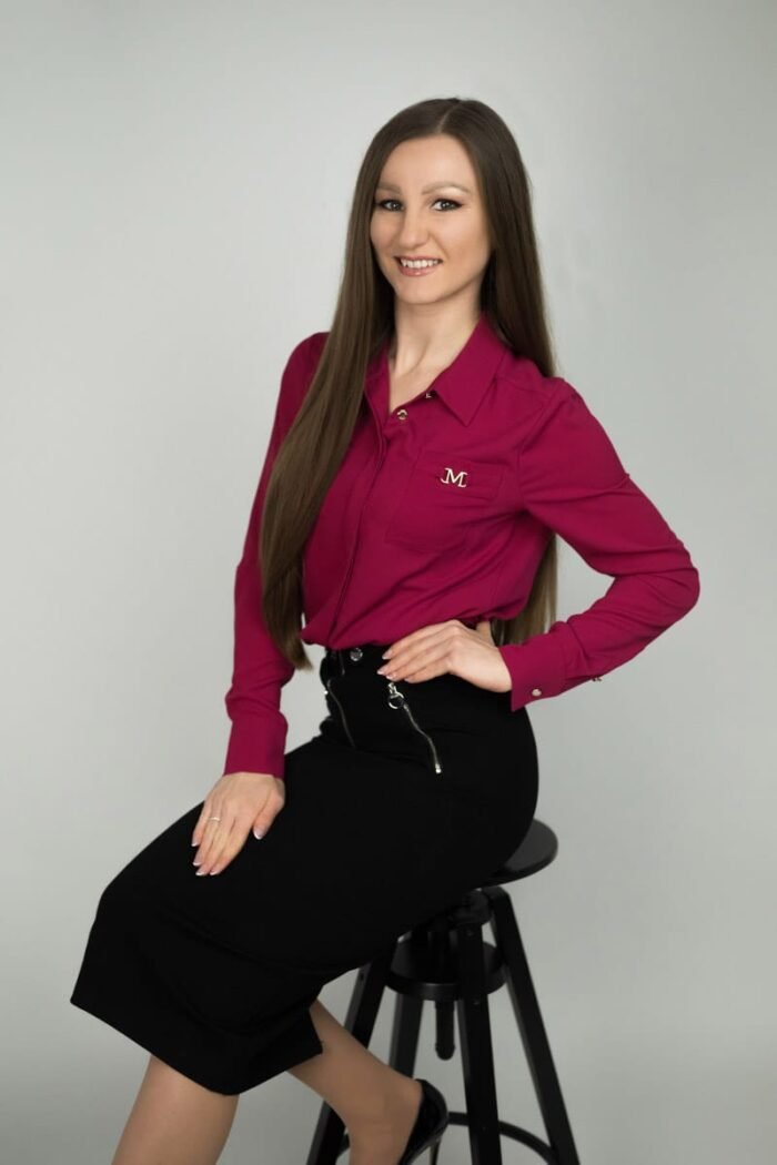 Egy mosolygós portréfotó világos stúdióban, ahol egy hosszú barna hajú nő pink inget és fekete szoknyát viselve ül egy széken, elegáns, de barátságos pózban.