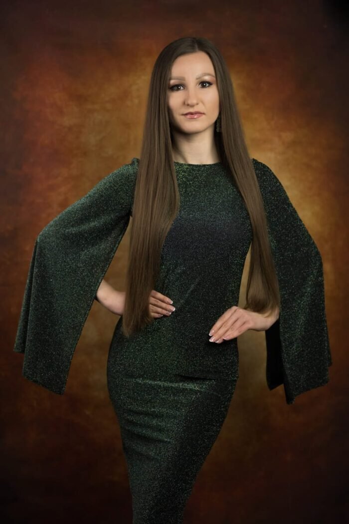 Egy elegáns portréfotó, amelyen egy hosszú barna hajú nő csillogó zöld ruhát viselve pózol, vintage stílusú, meleg tónusú háttér előtt.