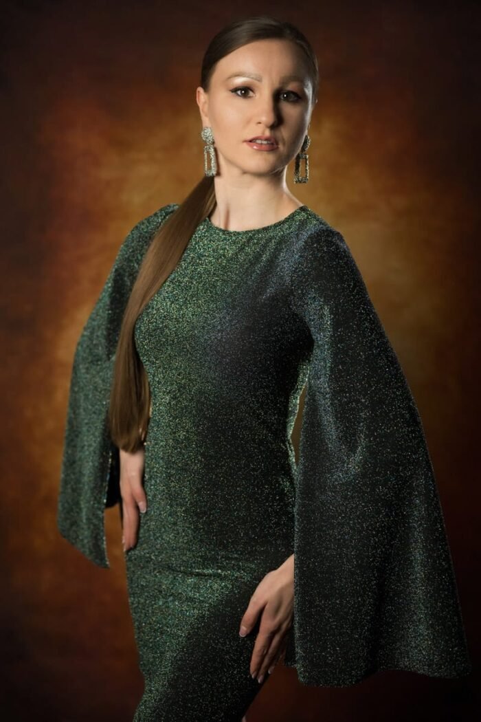 Egy elegáns portréfotó, amelyen egy hosszú barna hajú nő csillogó zöld ruhát és nagy, feltűnő fülbevalót viselve pózol vintage hangulatú, meleg tónusú háttér előtt.