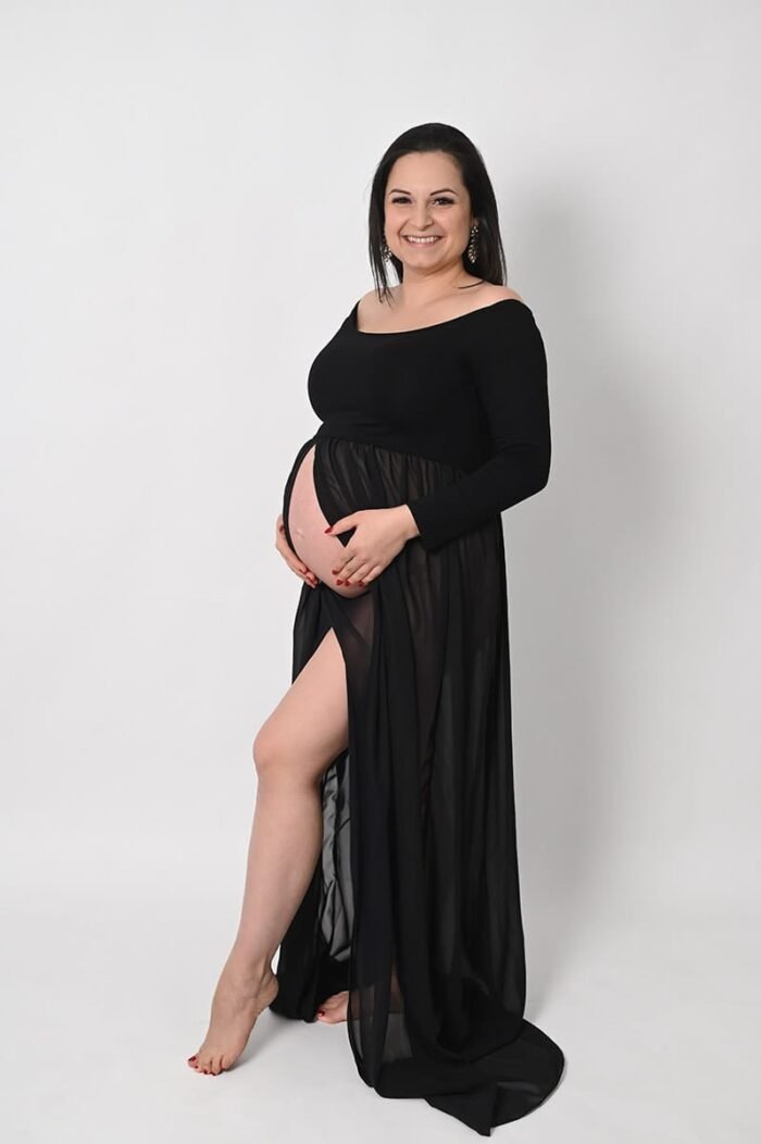 Terhes fotózás Debreceneben - Egy mosolygó kismama elegáns fekete ruhában, áttetsző szoknyarésszel áll a fehér háttér előtt, kiemelve várandós hasát.