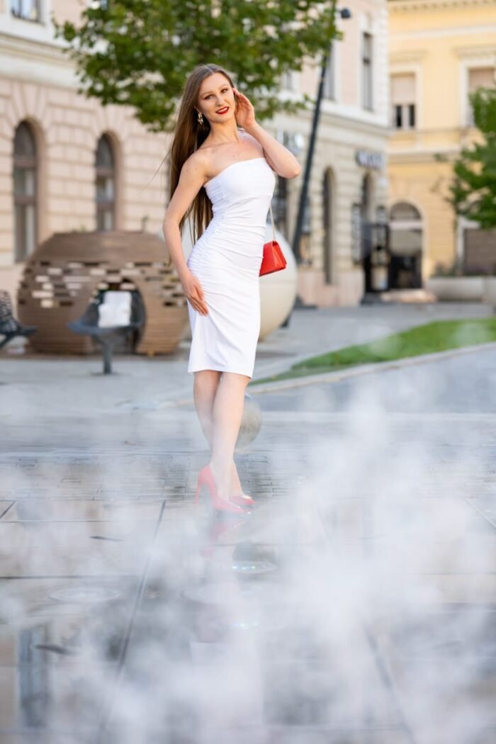 Fiatal nő fehér ruhában, piros magas sarkú cipőben és piros táskával pózol Debrecen belvárosában, miközben körülötte misztikus ködfátyol emelkedik.