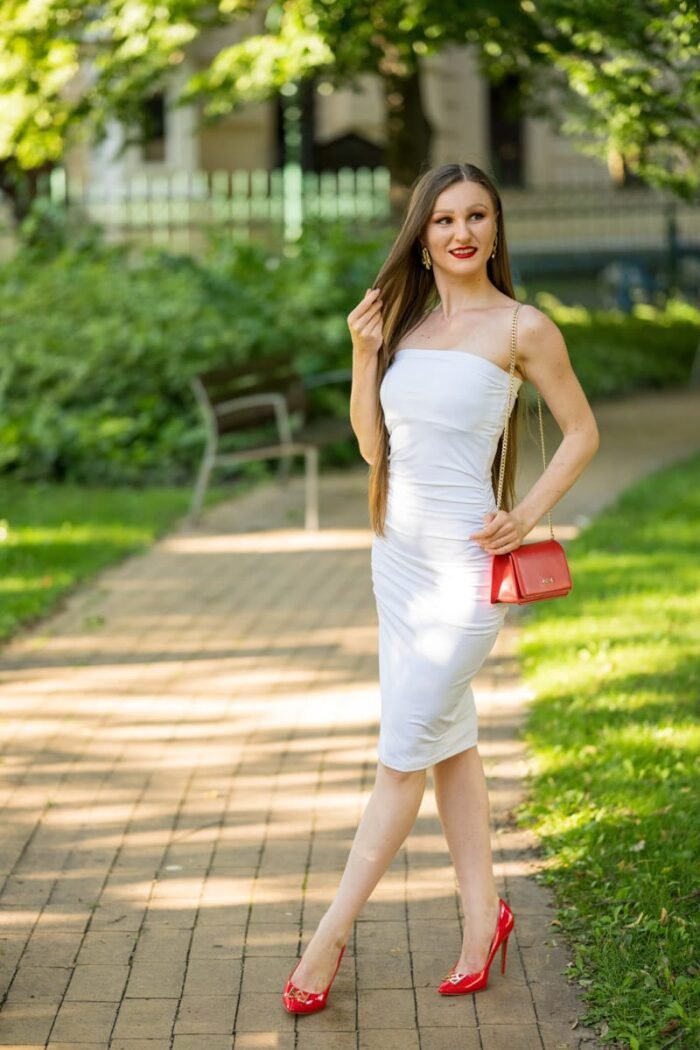 Fiatal nő elegáns fehér ruhában, piros magas sarkú cipőben és piros kistáskával sétál egy parkos környezetben Debrecenben, szabadtéri fotózás közben.