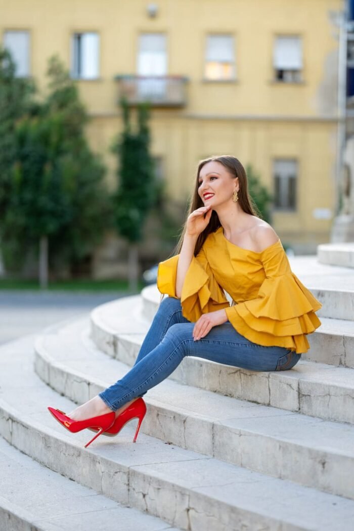Mosolygó nő sárga fodros felsőben és piros magassarkú cipőben, Debrecenben, lépcsőkön ülve, elegáns szabadtéri fotózás során.