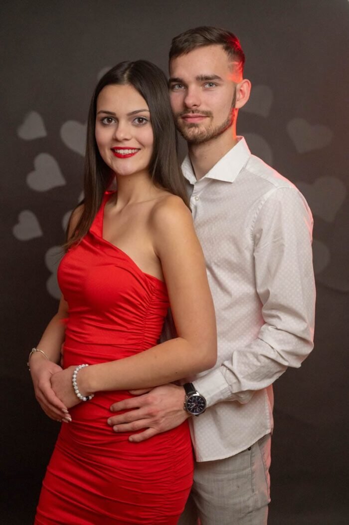 Fiatal pár Valentin-napi hangulatú stúdiófotón, a hölgy piros ruhában, a háttérben szív alakú dekorációval, romantikus stílusban.