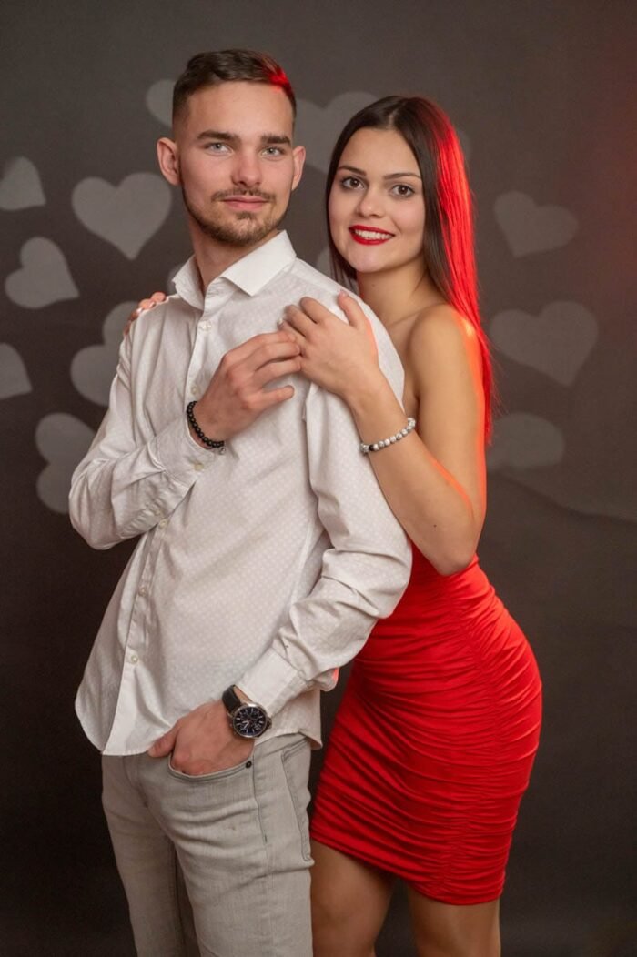 Fiatal pár Valentin-napi stúdiófotón, a férfi elegáns fehér ingben, a nő piros ruhában, háttérben szív alakú dekorációval.