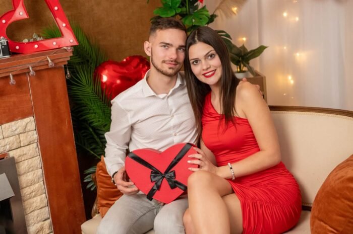 Fiatal pár ül egy kanapén, kezükben egy piros szív alakú ajándékdobozzal, Valentin-napi dekorációkkal a háttérben, romantikus hangulatban.