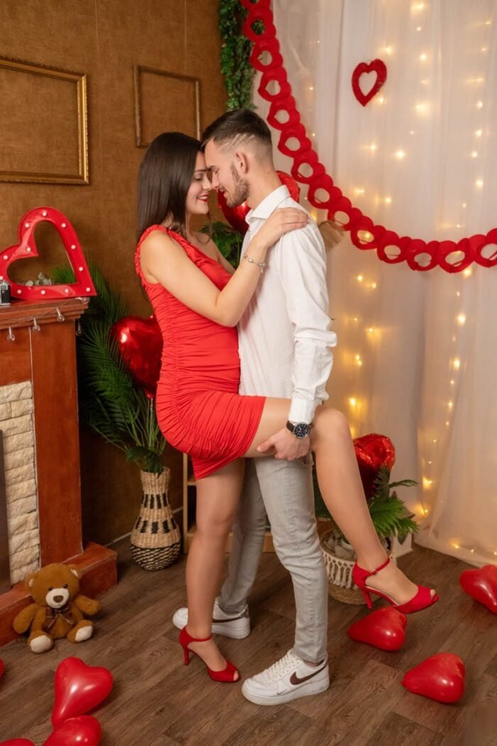 Fiatal pár Valentin-napi stúdiófotón, szenvedélyes pózban, romantikus dekorációval és piros szív alakú lufikkal körülvéve.