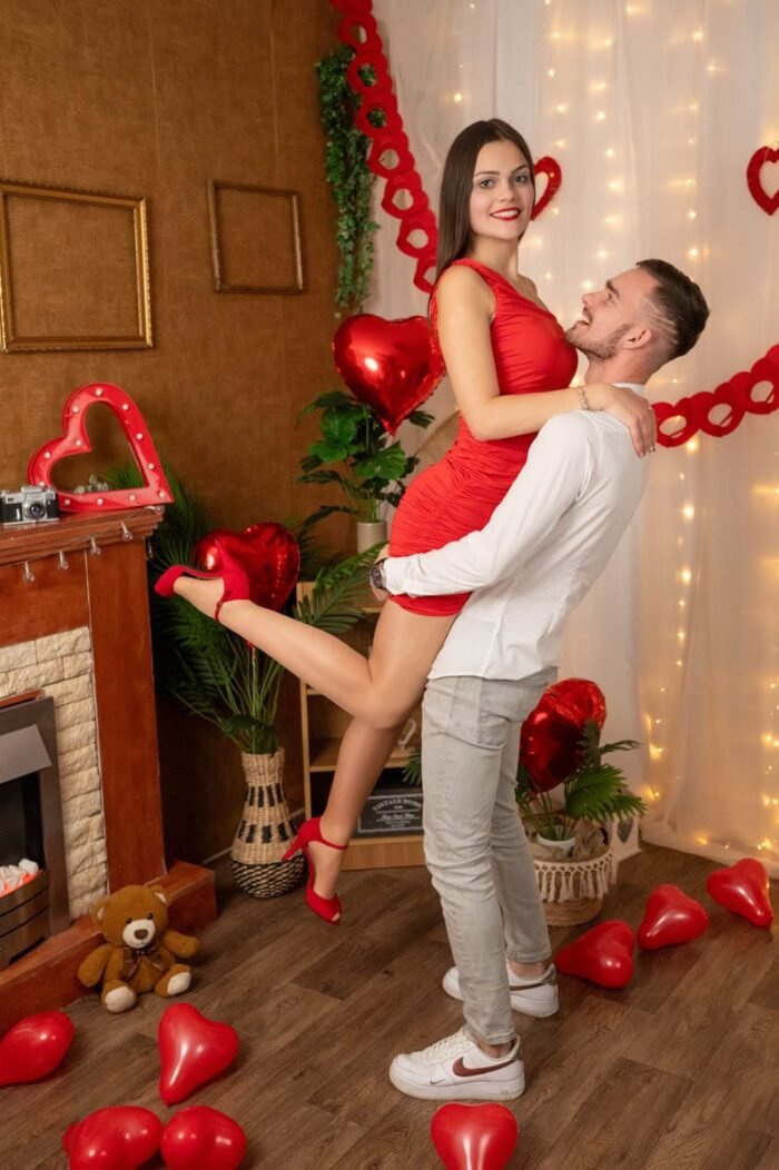 Fiatal pár Valentin-napi stúdiófotón, ahol a férfi felemeli a nőt, háttérben piros szív alakú dekorációk és romantikus hangulat.