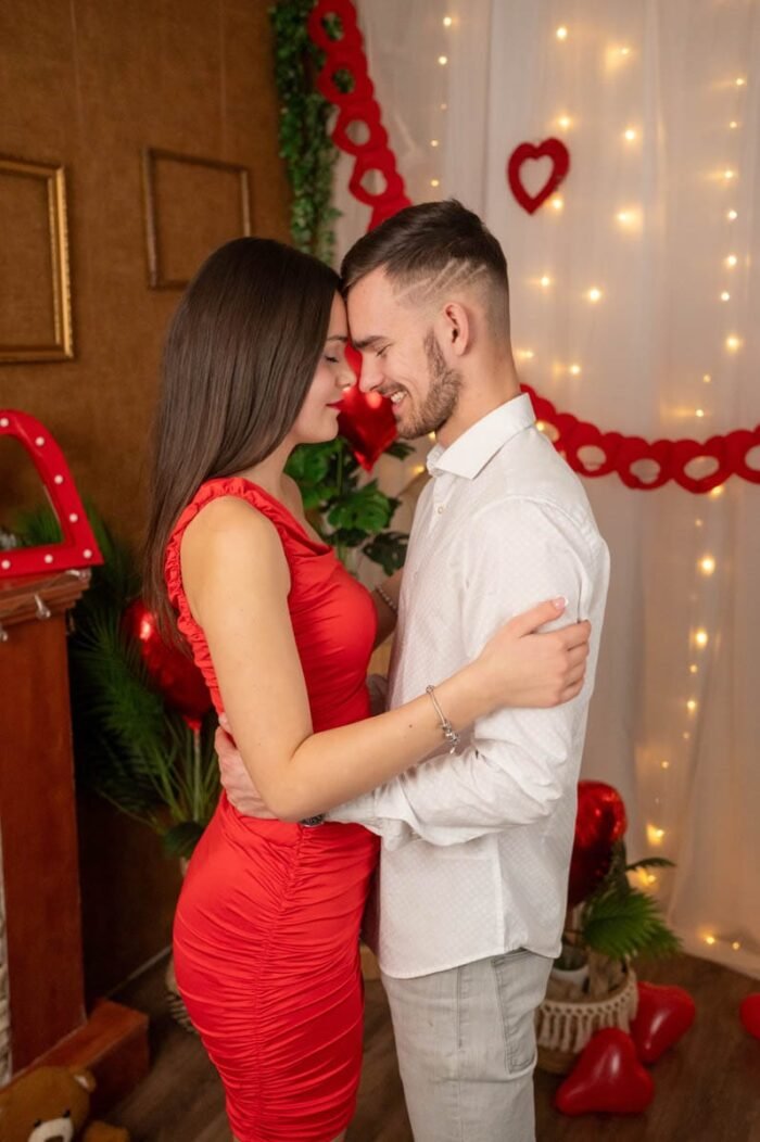 Fiatal pár meghitt ölelésben, mosollyal az arcukon, Valentin-napi stúdiófotón, romantikus dekorációk és piros szívek között.