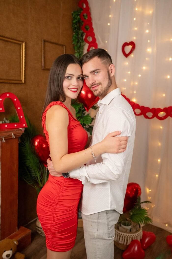 Fiatal pár Valentin-napi stúdiófotón, elegáns öltözetben, piros szív alakú dekorációk és romantikus hangulatú háttér előtt.