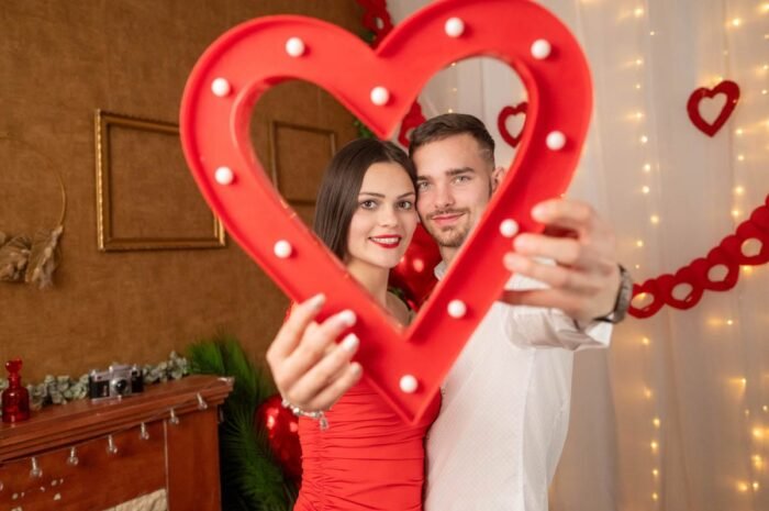 Fiatal pár mosolyog egy piros szív alakú dekoráció mögött, romantikus Valentin-napi stúdiófotón, hangulatos háttérrel.