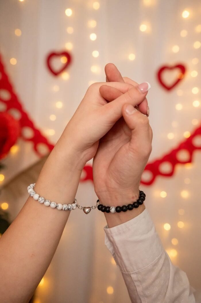 Szerelmes pár keze összeér, páros karkötőkkel, romantikus Valentin-napi dekoráció és hangulatvilágítás előtt.