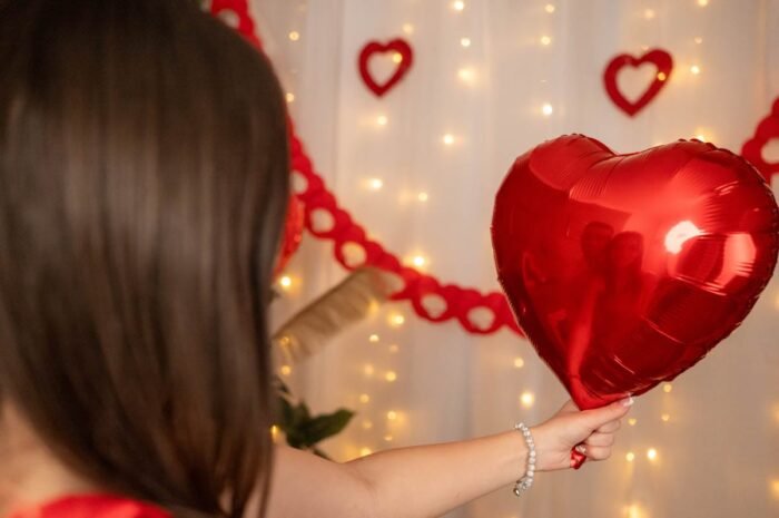 Piros szív alakú lufit tartó nő, amelyen visszatükröződik egy szerelmes pár, Valentin-napi romantikus dekoráció és meleg fények hátterében.