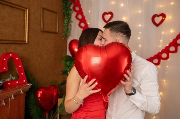 Egy pár Valentin-napi dekoráció előtt, akik egy nagy piros szív alakú léggömb mögött csókolóznak, miközben körülöttük szív alakú díszek láthatók.